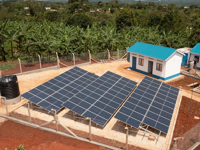 Three solar panels next to a few small houses with blue roofs, with a background of jungle foliage.