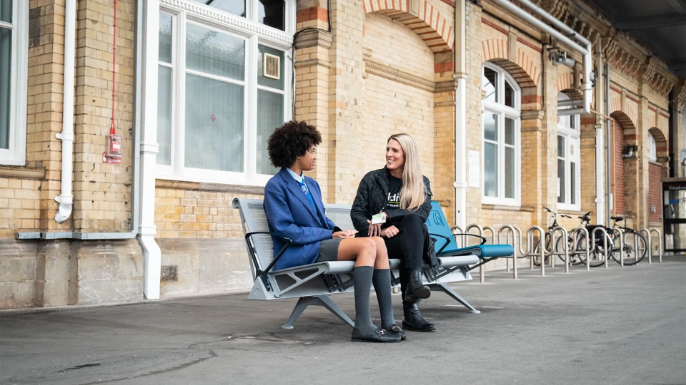 Child in school uniform sitting next to woman in train station platform chatting