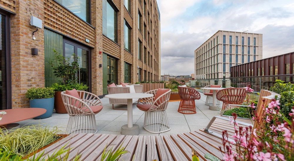 A spacious balcony at One Blossom Yard, UK, features chairs and a small table with potted plants. The skyline and distant mountains create a scenic backdrop against a cloudy afternoon sky.