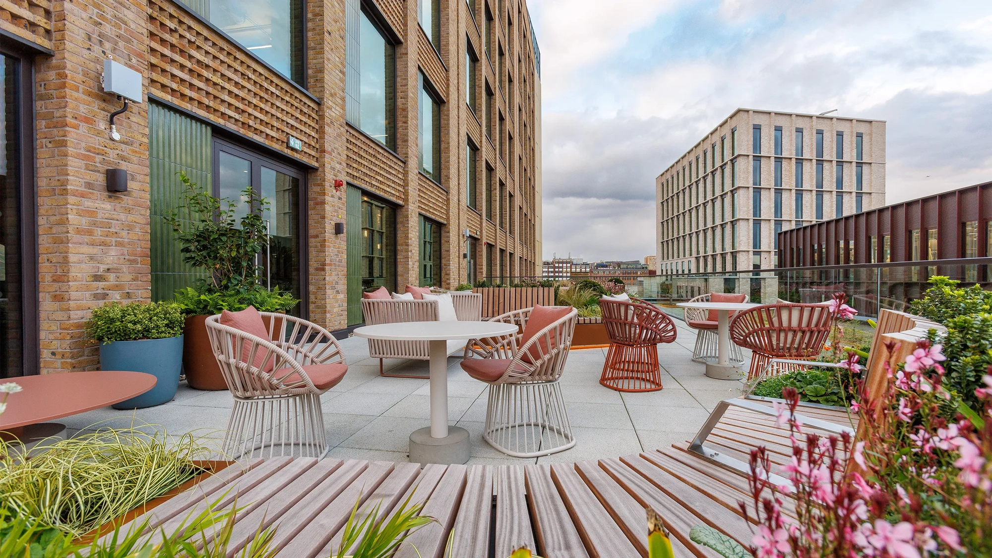 A spacious balcony at One Blossom Yard, UK, features chairs and a small table with potted plants. The skyline and distant mountains create a scenic backdrop against a cloudy afternoon sky.