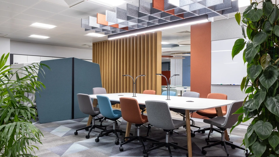 Turner & Townsend Bristol office. Plants in each corner with a meeting bank in the centre. There's colorful chairs around the table and a orange panel behind.