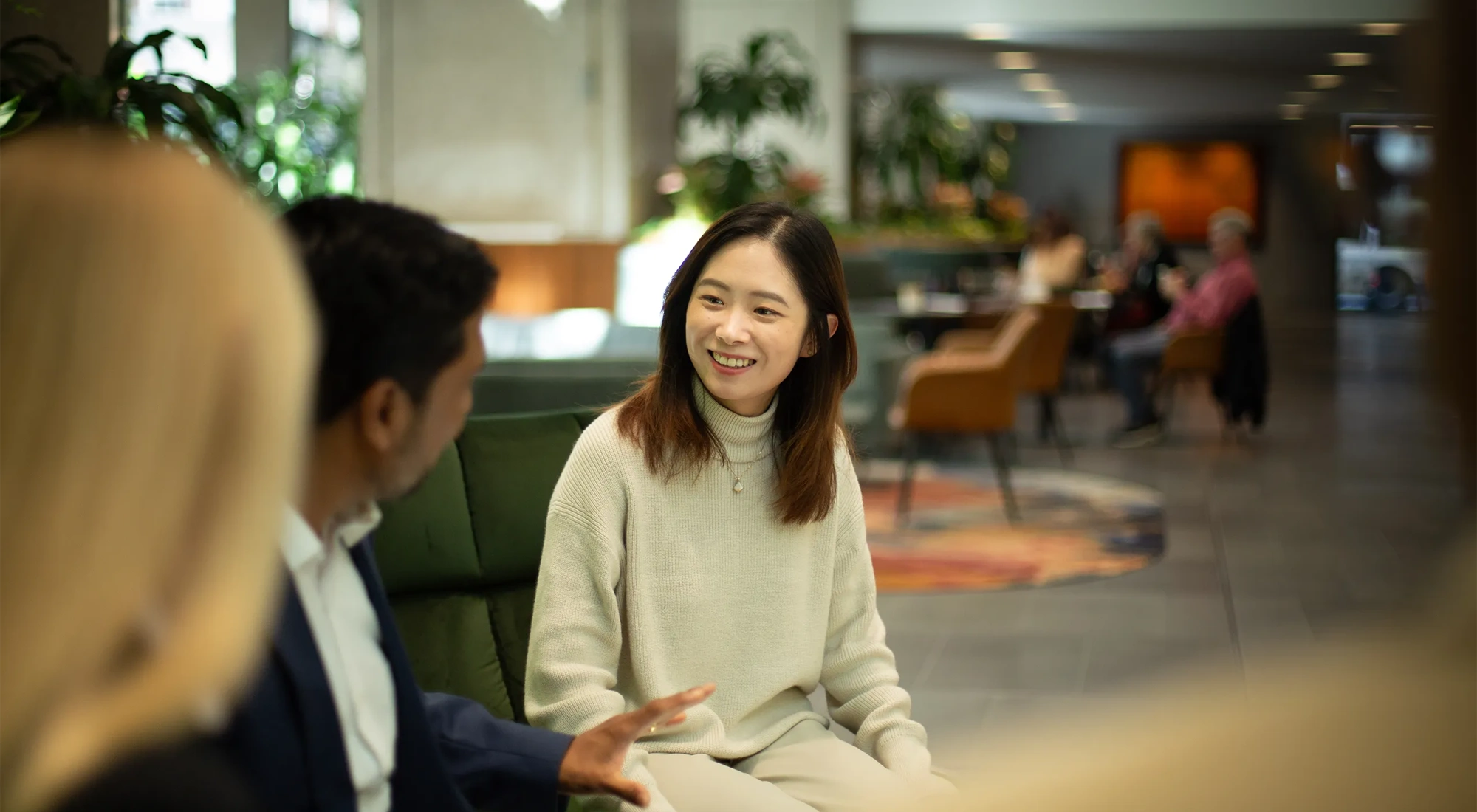 Three colleagues sitting together on dark green sofas having a conversation in a plant-filled office. Main focus is woman with short brown hair in neutral tone clothing.