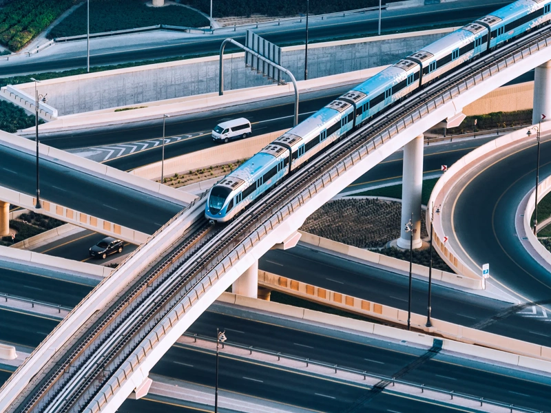 An elevated blue and white metro train travels along a curved, multi-level bridge over a network of highways. Several cars are visible on the roads below, navigating the complex system of interchanges. The image captures modern urban infrastructure, with smooth, sweeping curves and elevated tracks against a backdrop of landscaped areas and concrete structures