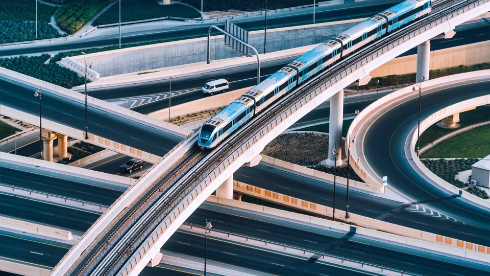 An elevated blue and white metro train travels along a curved, multi-level bridge over a network of highways. Several cars are visible on the roads below, navigating the complex system of interchanges. The image captures modern urban infrastructure, with smooth, sweeping curves and elevated tracks against a backdrop of landscaped areas and concrete structures