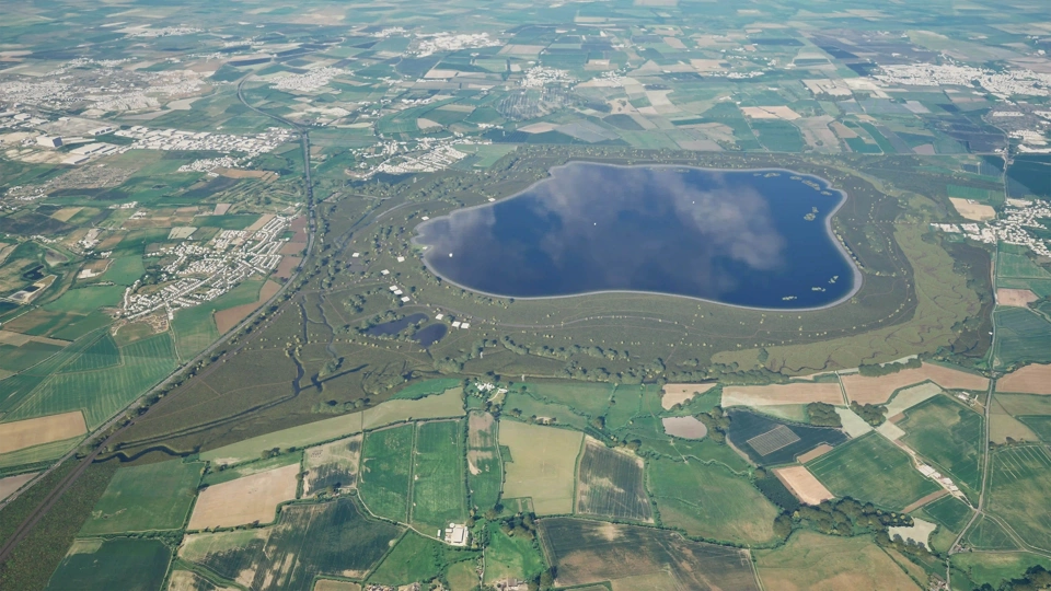 An aerial view of a large stone dam with a reservoir on the right side and a lush green landscape surrounding it. A small building is visible near the base of the dam, adjacent to a body of water and rocky terrain.