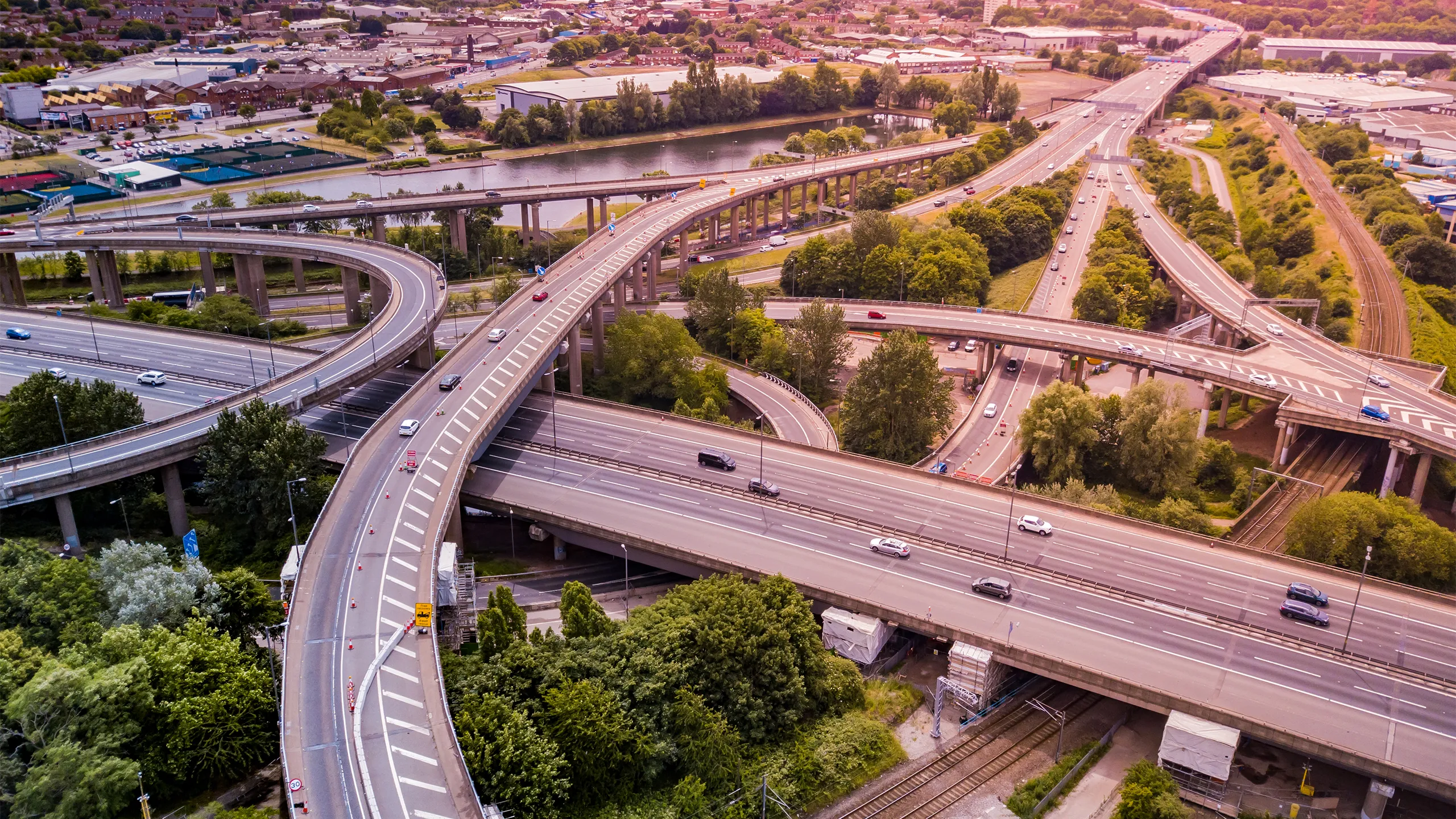 Aerial view of a complex motorway road junction with traffic moving. Cars, lorries, vans and a train can be seen travelling through a busy road interchange from above.