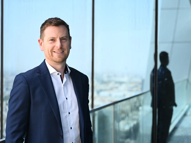 Alan Coary, Middle East data centre lead business suit standing on a balcony or near a large window, with a cityscape visible in the background.