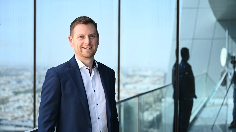 Alan Coary, Middle East data centre lead business suit standing on a balcony or near a large window, with a cityscape visible in the background.