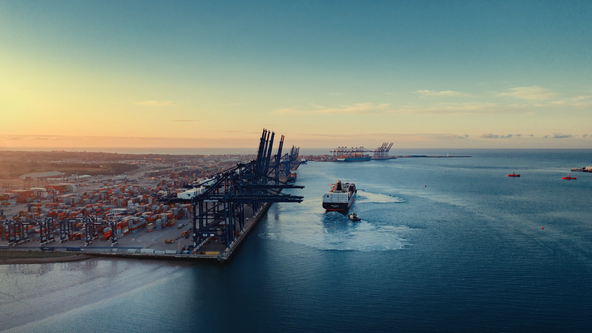 An aerial view of a large shipping port at sunset. The port is lined with numerous container cranes and stacks of shipping containers. A cargo ship is seen navigating out of the port towards the open sea, leaving a wake behind. The sky is clear with a gradient from light yellow to blue as the sun sets in the distance.