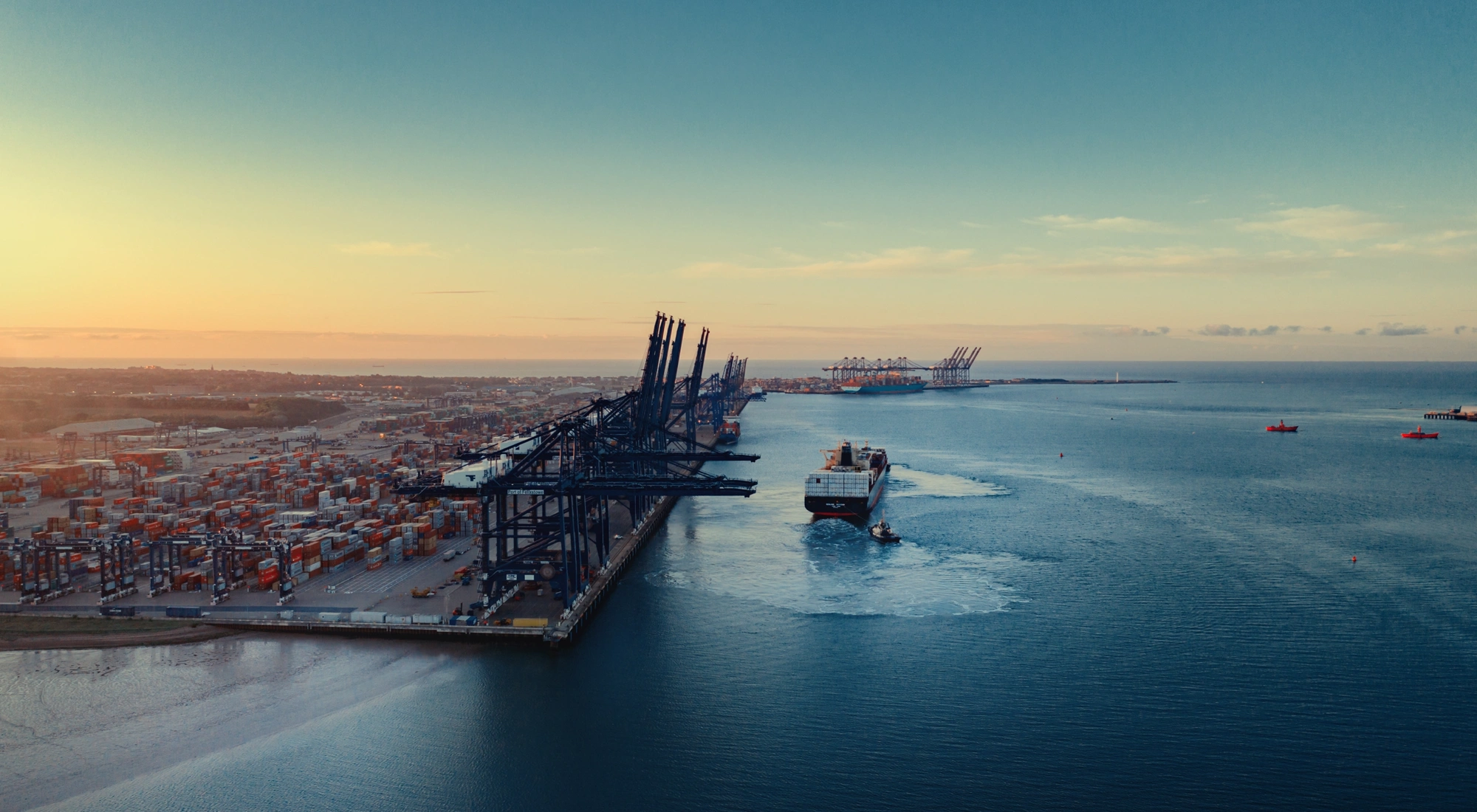 An aerial view of a large shipping port at sunset. The port is lined with numerous container cranes and stacks of shipping containers. A cargo ship is seen navigating out of the port towards the open sea, leaving a wake behind. The sky is clear with a gradient from light yellow to blue as the sun sets in the distance.