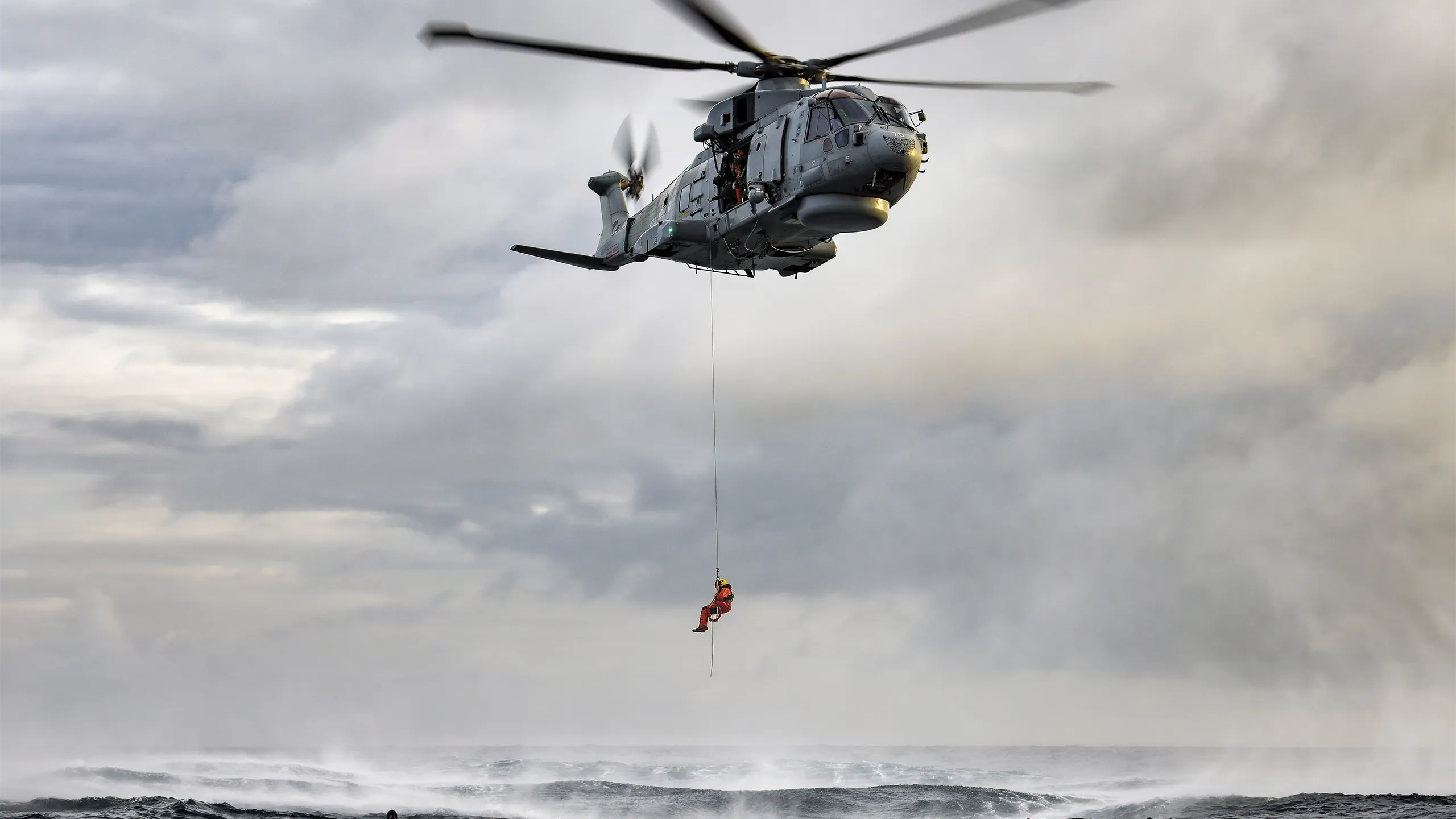 A crew member from a hovering military rescue helicopter is lowered by powered winch from a side door.