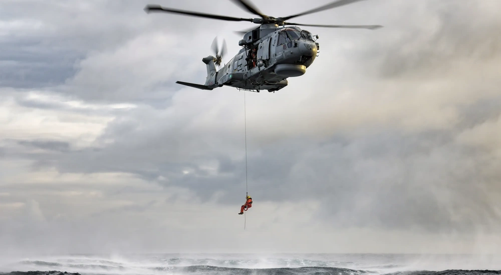 A crew member from a hovering military rescue helicopter is lowered by powered winch from a side door.
