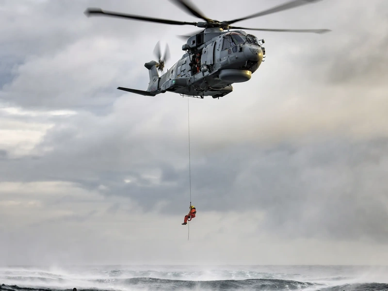 A crew member from a hovering military rescue helicopter is lowered by powered winch from a side door.
