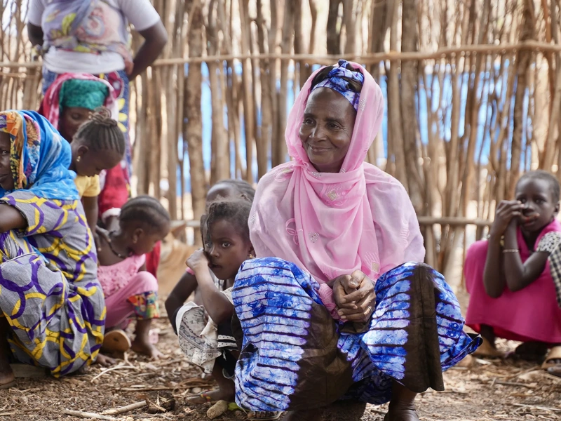 A group of women and children sit together outdoors in front of a wooden fence, wearing colorful patterned clothing.