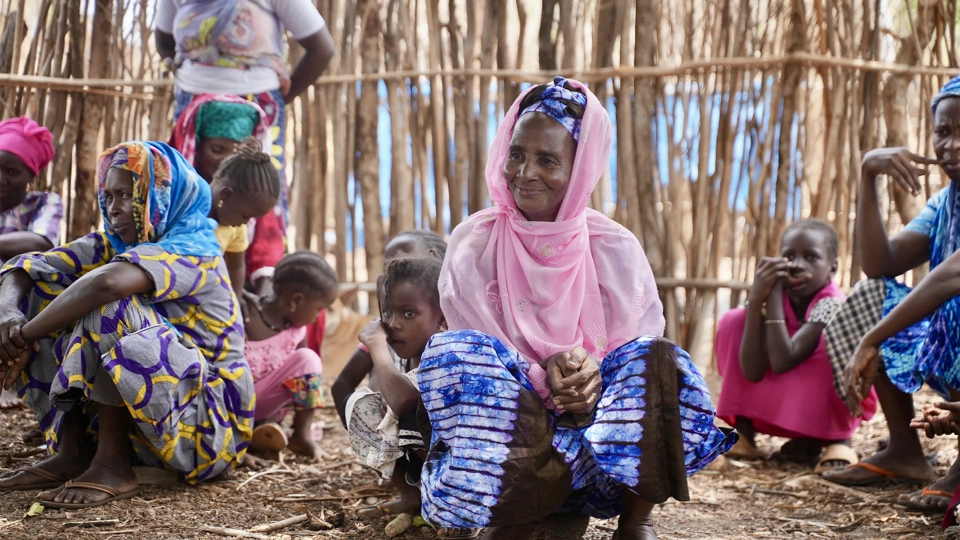 A group of women and children sit together outdoors in front of a wooden fence, wearing colorful patterned clothing.