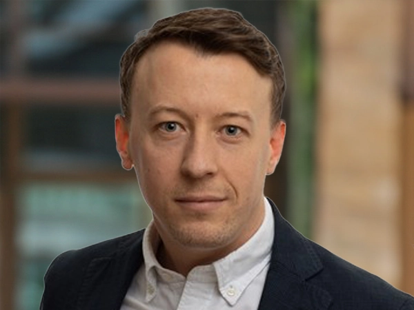 Oliver Molloy, Director, outside in front of a blurred brown background featuring a building and mix of natural and urban elements.