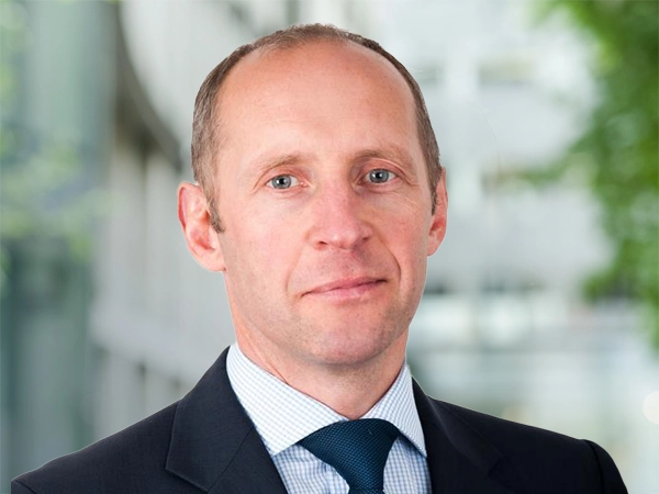 Gareth Morgan, Director, standing outside in front of an outdoor urban setting, featuring modern architectural elements with clean lines. There are hints of greenery, suggesting some trees or plants.