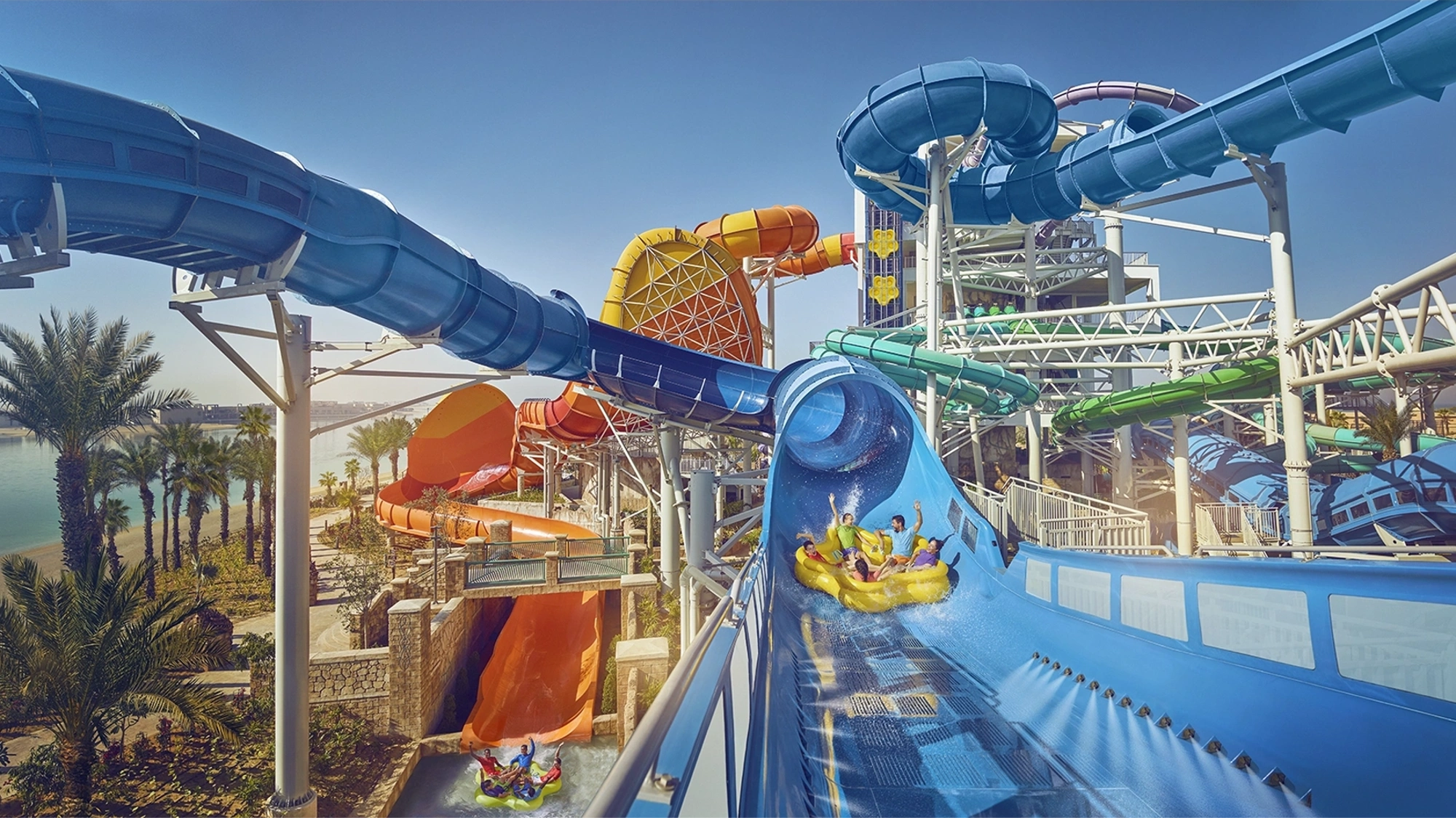 Big water park with lots of slides, featuring family on water float going down big blue water slide with heir arms raised in the air