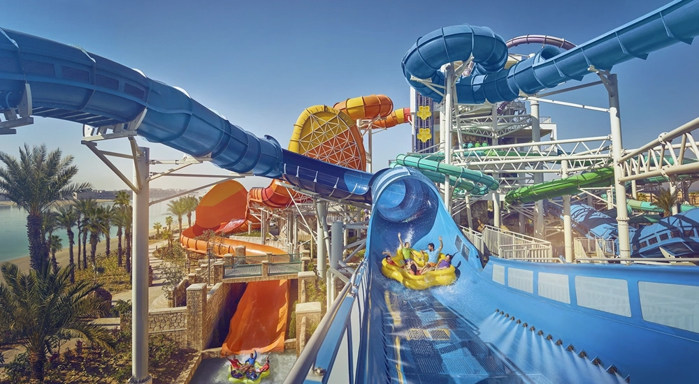 Big water park with lots of slides, featuring family on water float going down big blue water slide with heir arms raised in the air