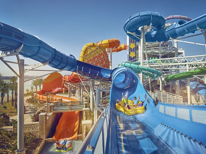 Big water park with lots of slides, featuring family on water float going down big blue water slide with heir arms raised in the air