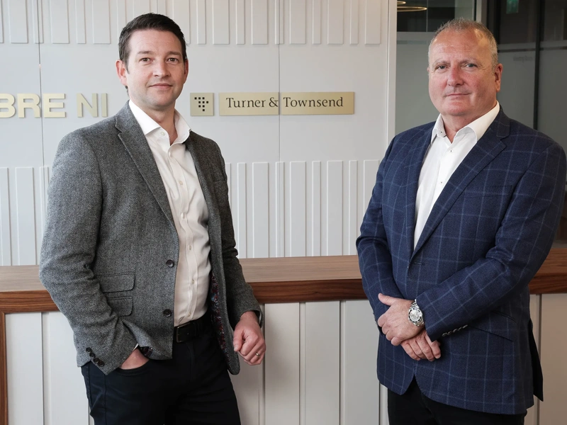 Two business men in suits, standing in front of grey back ground with gold CBRE and Turner & Townsend logo.