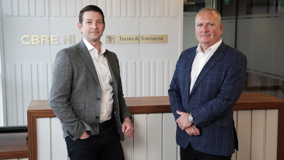 Two business men in suits, standing in front of grey back ground with gold CBRE and Turner & Townsend logo.