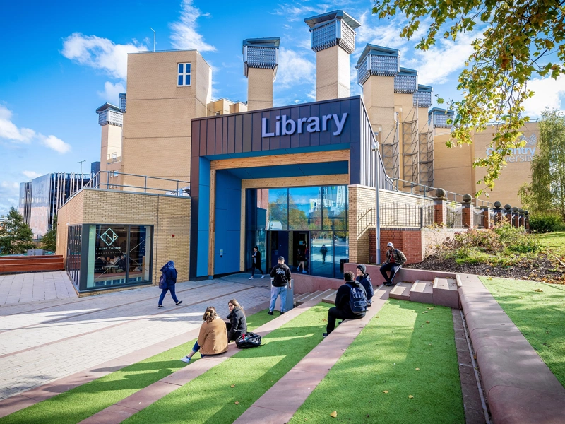 Students sitting on a lawn and stairs outside a library building