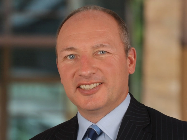  James Hamilton, Director, Global Host Cities and Sports Venues, outside in front of a blurred brown background featuring a building and mix of natural and urban elements.