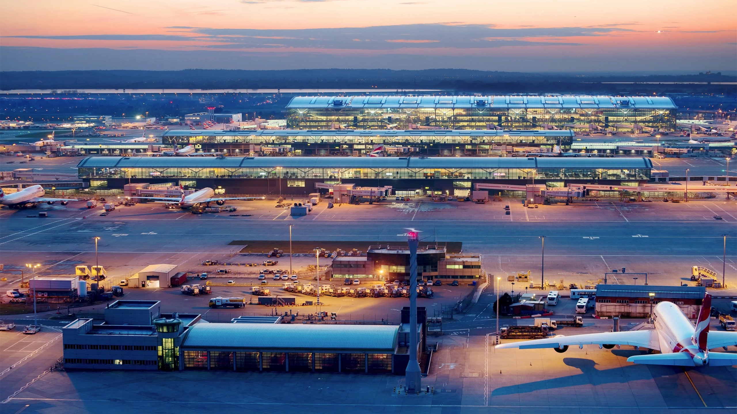 Heathrow airport featuring an airplane, bright lights in the building and orange sunset in the background