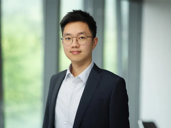 Portrait of Isaac Hou, Construction Economist, standing infront of windows in office with faint trees in the background smiling.