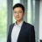 Portrait of Isaac Hou, Construction Economist, standing infront of windows in office with faint trees in the background smiling.
