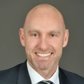 Portrait of Mark Utting, Managing Director, Switzerland, smiling in a suit against dark grey background.