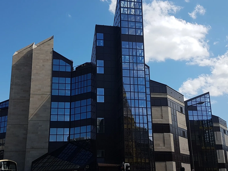 modern building with a distinctive architectural style. The building features a mix of concrete and dark glass facades, with several angular, geometric shapes and pointed rooflines. The structure appears to have multiple sections, including a central tower with a glass facade that reflects the sky and surrounding structures. The sky is clear with a few scattered clouds, providing a bright contrast to the dark glass of the building.