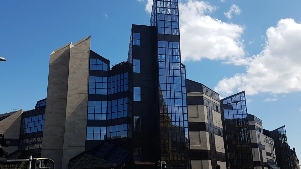 modern building with a distinctive architectural style. The building features a mix of concrete and dark glass facades, with several angular, geometric shapes and pointed rooflines. The structure appears to have multiple sections, including a central tower with a glass facade that reflects the sky and surrounding structures. The sky is clear with a few scattered clouds, providing a bright contrast to the dark glass of the building.