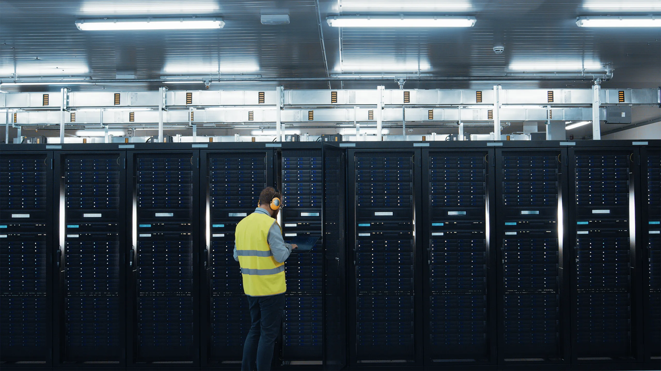 IT Engineer in High Visibility Vest is Working on Laptop in Data Center while Standing Before Server Rack. Running Diagnostics or Doing Maintenance Work.