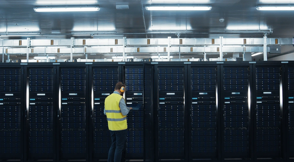 IT Engineer in High Visibility Vest is Working on Laptop in Data Center while Standing Before Server Rack. Running Diagnostics or Doing Maintenance Work.