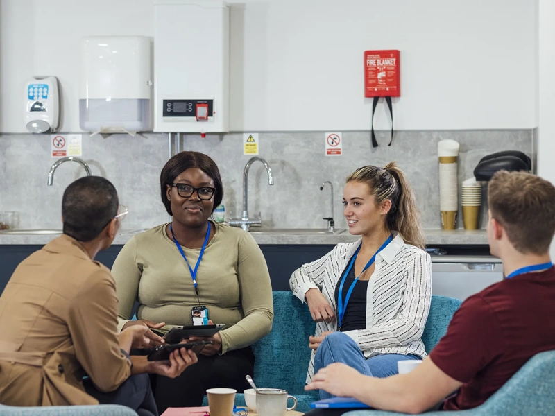 A group of healthcare professionals sitting in chairs around a wooden table in a staff common room in a hospital in Newcastle upon Tyne, North East England. They are talking and laughing while taking a break from working.