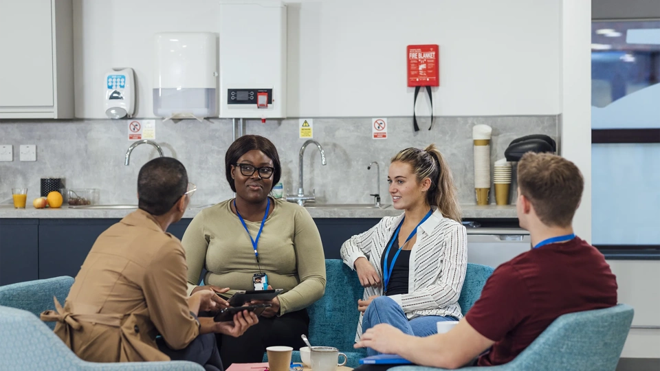 A group of healthcare professionals sitting in chairs around a wooden table in a staff common room in a hospital in Newcastle upon Tyne, North East England. They are talking and laughing while taking a break from working.
