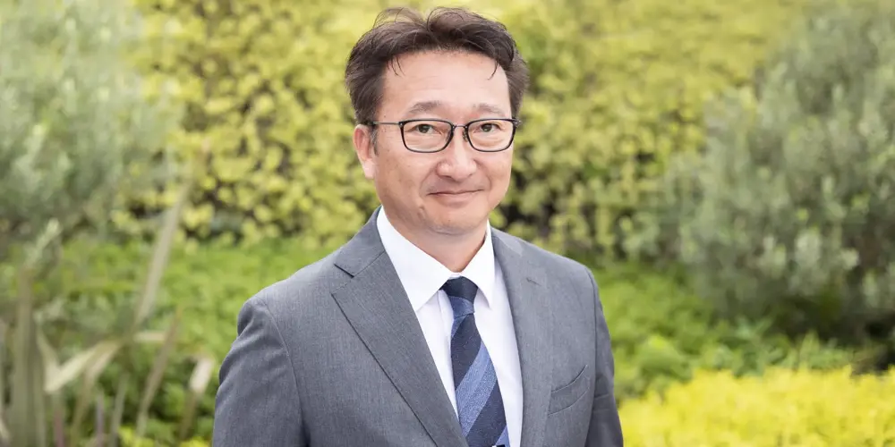 Portrait of Toshio Kosuge, director of clean energy and transmissions in Japan, standing outside against a green background