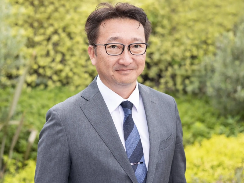 Portrait of Toshio Kosuge, director of clean energy and transmissions in Japan, standing outside against a green background
