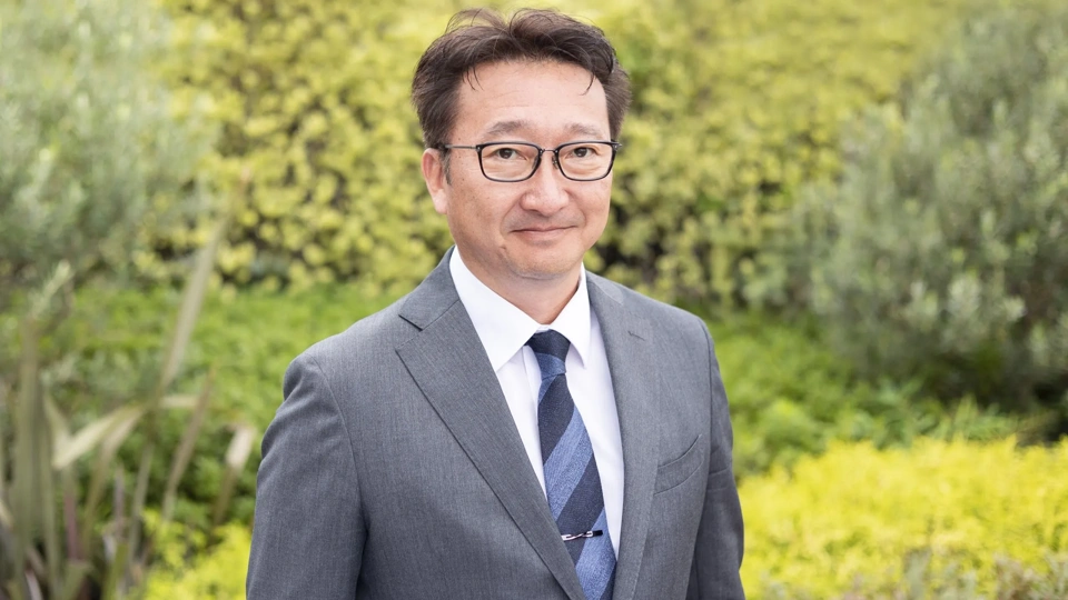 Portrait of Toshio Kosuge, director of clean energy and transmissions in Japan, standing outside against a green background