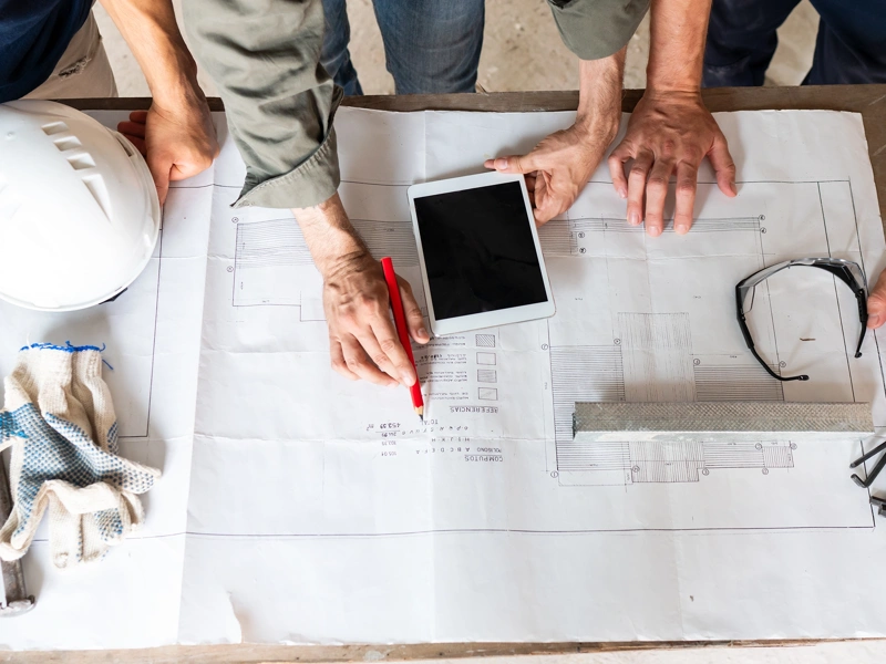 Three work people reading building plans on a table, includes iPad and construction work hat and safety glasses
