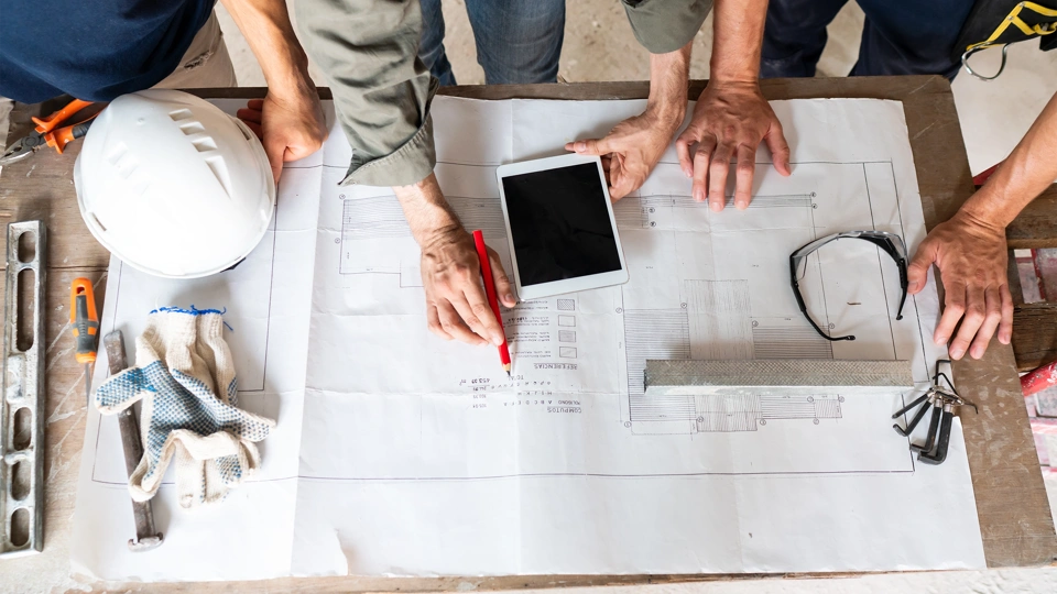 Three work people reading building plans on a table, includes iPad and construction work hat and safety glasses