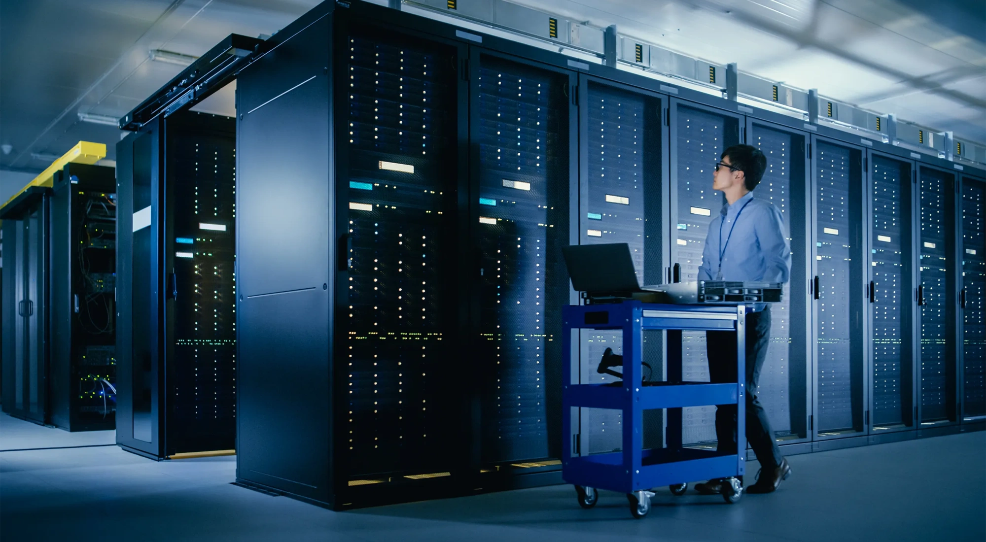 In the Modern Data Center: IT Engineer Doing Maintenance and Diagnostics. Pushes Cart alongside Server Racks, Ready to Install New Hardware for Planned System Update, Equipment Replacement.