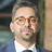 Satch Bhattacharjee, Key Client Lead, Australia, outside in front of a blurred brown background featuring a building and mix of natural and urban elements.