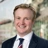 Portrait of George Highfield, Real Estate Advisory, Data & Digital – Principal Consultant, standing outside in front of buildings featuring an urban cityscape with a mix of architectural styles. The scene includes some windows and red features, adding a touch of color.
