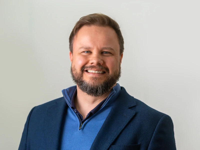 Portrait of Jason Byrne, Director for Integrated Advisory, UK at Turner & Townsend, standing against white background in blue suit, smiling.