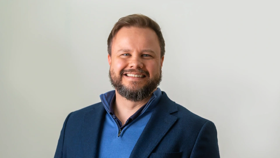 Portrait of Jason Byrne, Director for Integrated Advisory, UK at Turner & Townsend, standing against white background in blue suit, smiling.
