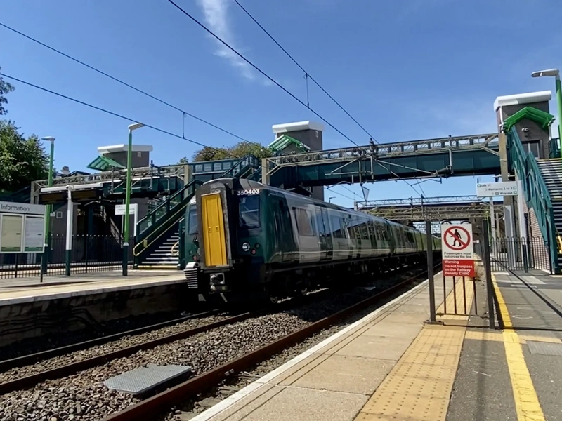 train station platform with a modern train passing through. Overhead electrified lines are visible, and there is a footbridge connecting both sides of the station. The station has green and white signage, and the platform area includes a yellow tactile paving strip for safety. Stairs with green railings lead up to the footbridge on either side, and a "No Entry" sign is visible near the platform edge. The sky is clear and blue, indicating a sunny day.