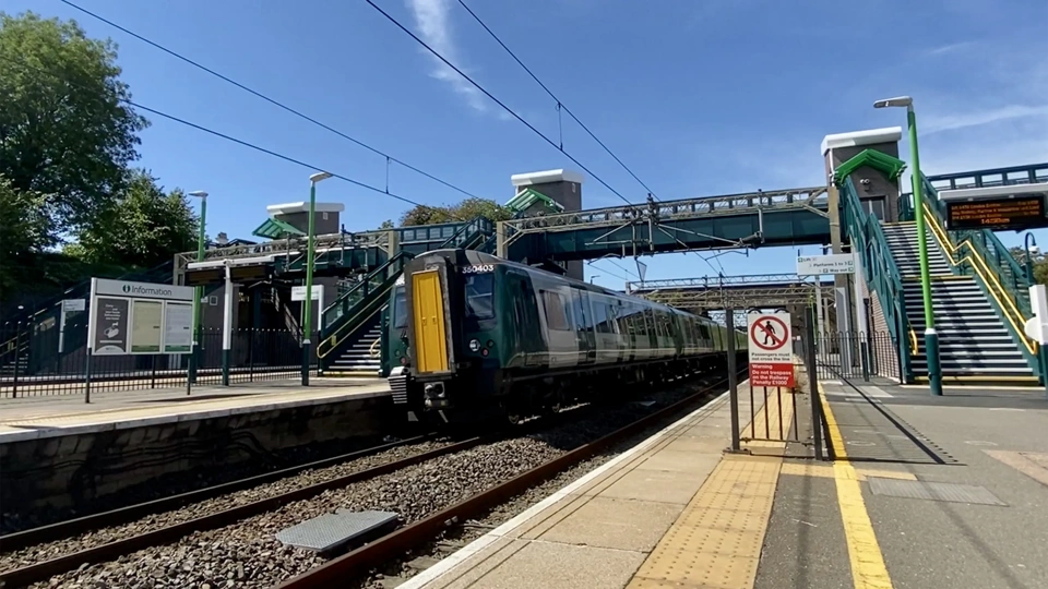 train station platform with a modern train passing through. Overhead electrified lines are visible, and there is a footbridge connecting both sides of the station. The station has green and white signage, and the platform area includes a yellow tactile paving strip for safety. Stairs with green railings lead up to the footbridge on either side, and a "No Entry" sign is visible near the platform edge. The sky is clear and blue, indicating a sunny day.